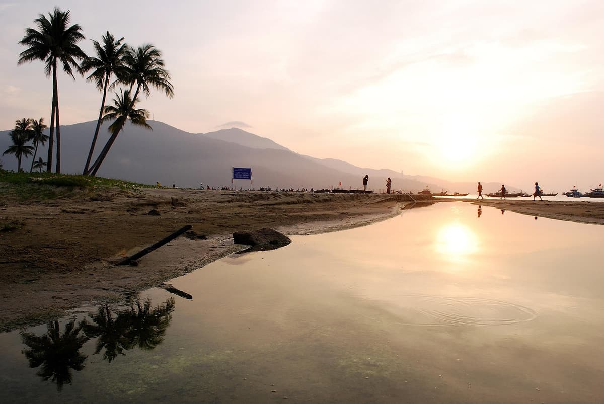 A long stretch of white sand and gentle waves at My Khe Beach in Da Nang with palm trees and high-rise hotels behind the coastal road