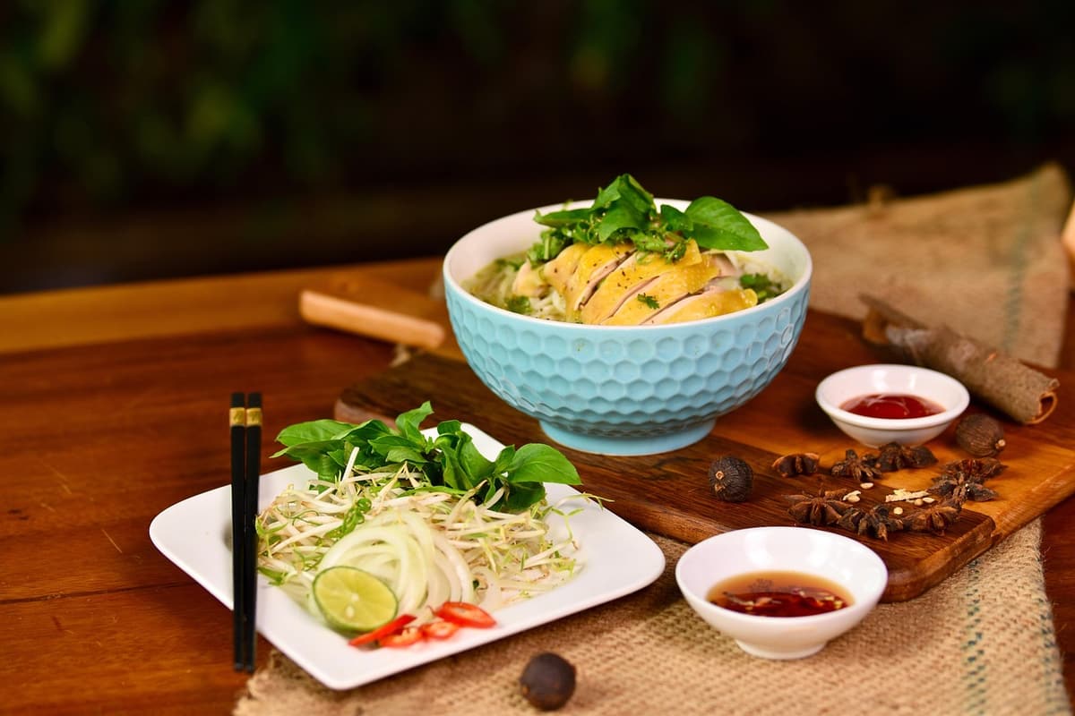A bowl of pho noodle soup in front of a Vietnamese street food stall with herbs, lime, and chili on the side and a small child holding chopsticks